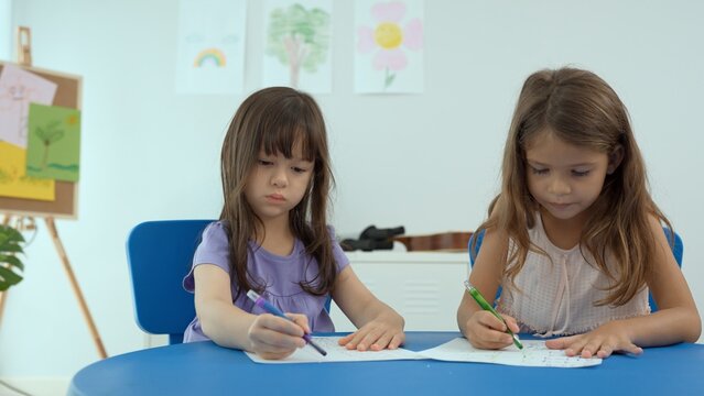 Woman teacher teaching little girl to paint color book on the table in classroom,kindergarten education school.Multi-ethnic preschool teacher and students in classroom.Kindergarten and study concept.