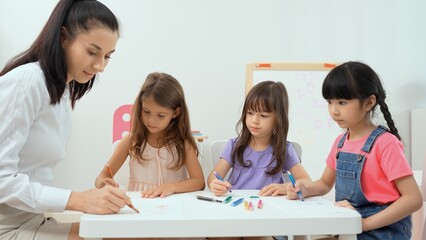 Woman teacher teaching little girl to paint color book on the table in classroom,kindergarten...