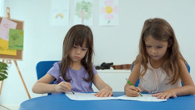 Woman teacher teaching little girl to paint color book on the table in classroom,kindergarten education school.Multi-ethnic preschool teacher and students in classroom.Kindergarten and study concept.