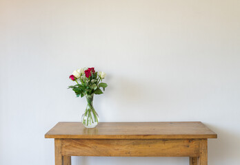 Red and cream roses in glass vase on oak side table against textured beige wall with copy space