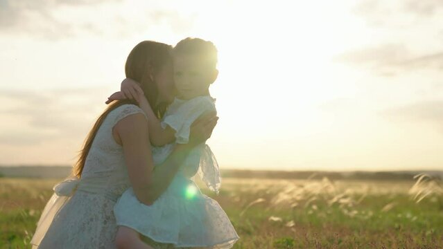 Happy family on walk in field in nature. Child, daughter hugs her mother in summer park in sun. Carefree childhood, joyful girl smiling, hugging mother and kid. Parent, kid play together at sunset