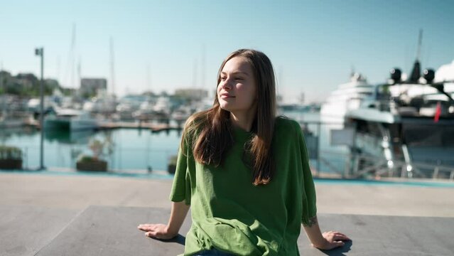 Relaxed blonde woman wearing green t-shirt sitting on the seafront near yachts outdoors
