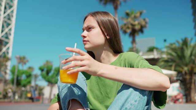Sunny blonde woman wearing green t-shirt looking around and drinking orange juice outdoors