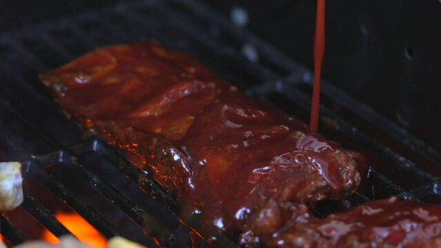 This video shows bbq sauce being poured on ribs in slow motion as they cook by fire on a flaming grill.