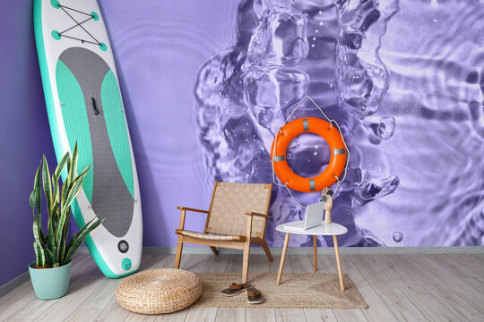 Interior of room with armchair, table, lifebuoy ring and board for sup surfing near wall with print of clear water