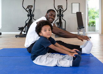 Young curly black hair boy stretching with his father after finish exercise routine on yoga mat. Cardio machines are on the background at the gym.