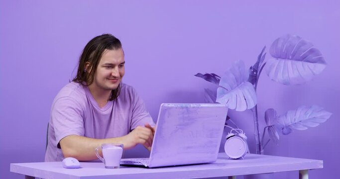 Serious young man with long hair works hard and gets angry, but successfully finishes the project, so he smiles and closes the laptop. Everything is painted purple