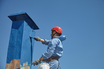 Naklejka premium An employee of the Industrial Mountaineering Service paints the chimney on the roof with a spray gun. Professional climber in uniform, helmet and with seat belts. Risky work. Extreme activity.
