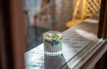 A glass of Iced matcha latte green tea with ice and milk on a wooden bar over a cafe glass window reflex at a Cafe coffee shop. Cold brew refreshment summer drink with copy space. Selective focus