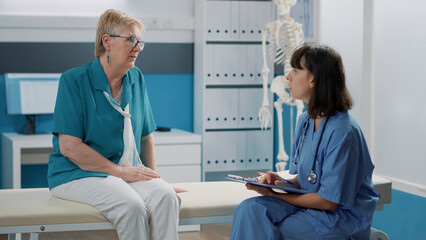 Female nurse taking notes on files after examination with old patient, having conversation about mechanical disorders in cabinet. Medical assistant discussing health care with elder woman.