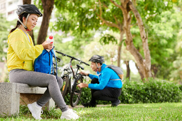 Asian young woman in bicycle helmet drinking water when boyfriend checking their bicycles in background