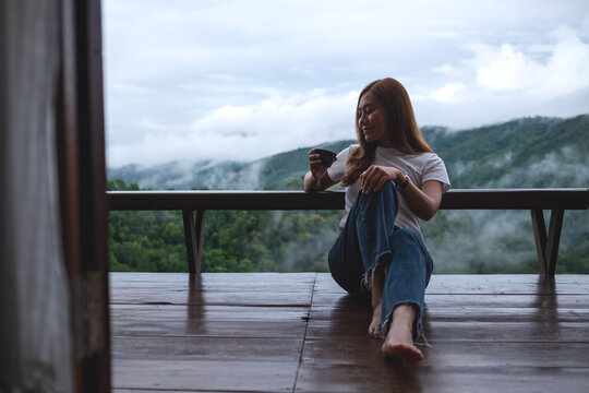 Portrait image of a woman drinking hot coffee while sitting on wooden balcony with a beautiful mountains and nature view on foggy day