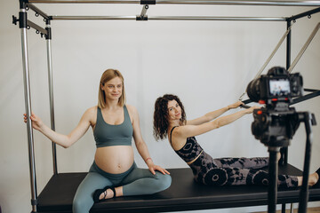 Fototapeta premium pregnant woman pilates reformer roll up cadillac exercise with personal trainer. Record a video blog