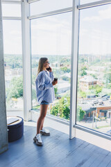 Fototapeta premium Modern woman freelancer talking on the phone while standing at a large window and drinking coffee. The concept of a modern successful woman. Young girl in an open office