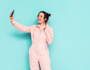 Young beautiful smiling female in trendy summer blue overalls. Sexy carefree woman posing near wall in studio. Positive brunette model looking at cellphone screen. Holding smartphone and using apps