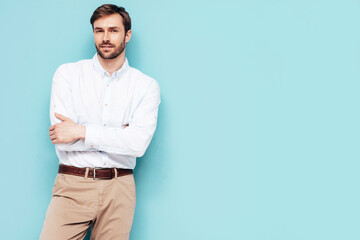 Portrait of handsome smiling model. Sexy stylish man dressed in shirt and trousers. Fashion hipster male posing near blue wall in studio. Crossed arms