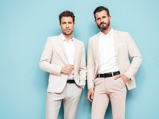 Portrait of two handsome confident stylish hipster lambersexual models. Sexy modern men dressed in white elegant suit. Fashion male posing in studio near blue wall
