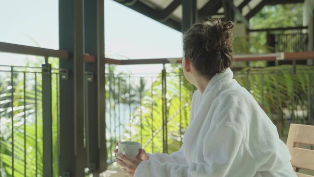 Female looks around hotel resort while sitting on balcony with coffee or tea. Wearing white hotel robe relaxing looking at tropical view with palm trees and beaches smiling and joyful in vacation mode