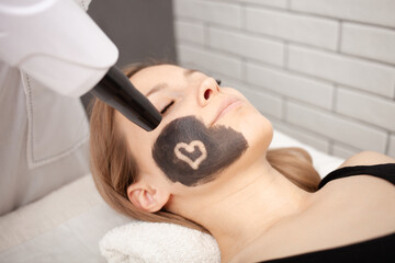 Obraz na płótnie Canvas cosmetologist does procedure of carbon peeling of face. Black mask, laser skin cleansing. Modern treatments. Cuts heart.