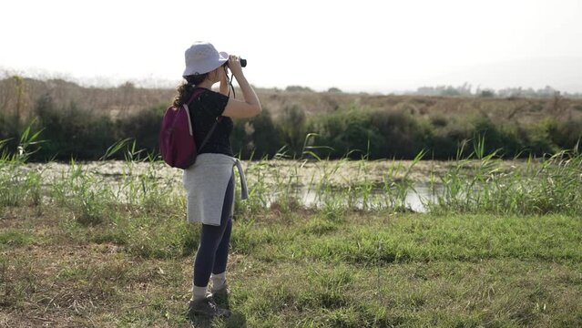 Female birdwatcher or nature enthusiast observing through binoculars outdoors. Young woman standing in natural reserve and watching animals from afar. Wearing backpack and hat during hot day