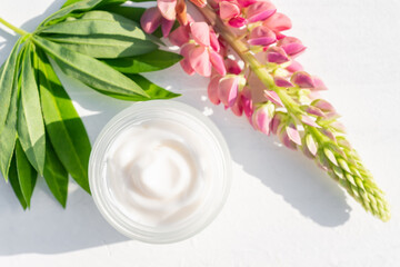 Unbranded white cosmetic cream ina open jar and bright lupine flowers on white background. Mockup, template. Container for body lotion, moisturizer cream.