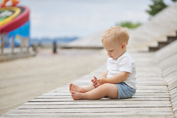 A cute baby boy is sitting on the ground at the playground outdoors. Adorable baby learning to walk. Healthy physical development. Funny child. Authentic lifestyle, happy childhood.