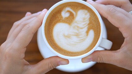 A mug of fragrant coffee held by female hands. Woman drinking morning sappuccino coffee from a white mug at a wooden table, top view. Coffee drink invigorates the body and spirit.