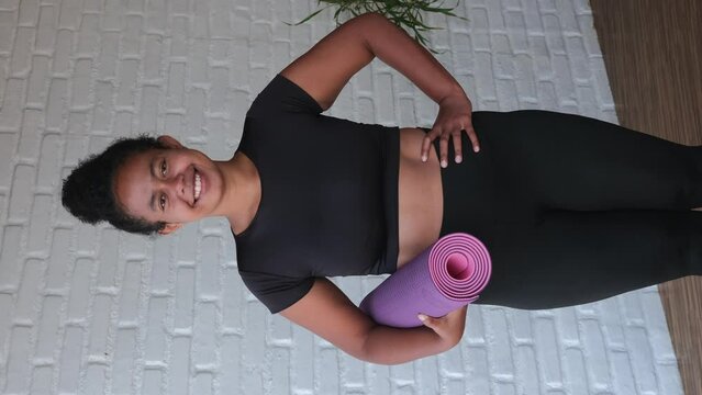 A dark-skinned woman in a rolled-up lilac yoga mat stands standing, smiling sweetly. Her smile is impressive. Metis woman is ready to practice yoga. Zambo model. vertical video.