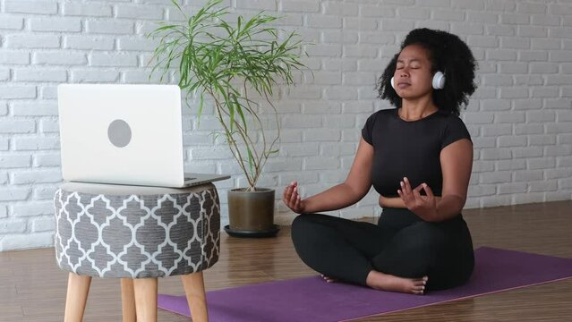 A woman in headphones in front of a laptop does yoga online. In a spacious room there is green vegetation, a stool with soft upholstery and computer equipment. A meditates on a purple mat.