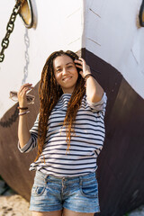 Fototapeta premium a girl with a dreadlocked hairstyle poses on the beach near a ship in summer, a sunny day, dressed in a T-shirt and denim shorts