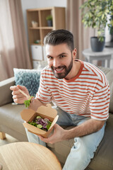 consumption, delivery and people concept - smiling man with fork and knife eating takeaway food at home