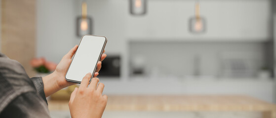 A mockup screen of a smartphone in a woman's hand over blurred modern white kitchen room - obrazy, fototapety, plakaty