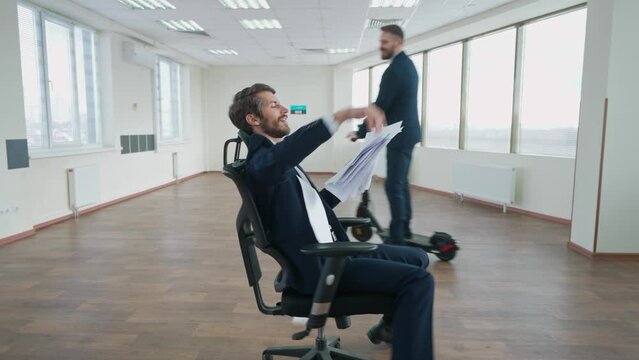 Office workers rejoice at successful completion of the project, man throws documents sitting on a chair, a colleague rides a scooter around the office space, flying papers, positive emotions.