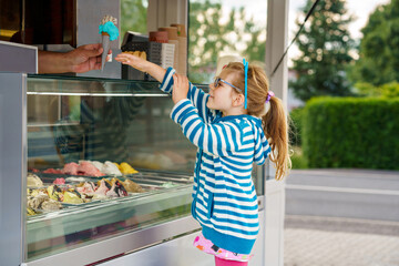 Cute little toddler girl choosing and buying ice cream in outdoor stand cafe. Happy preschool child...