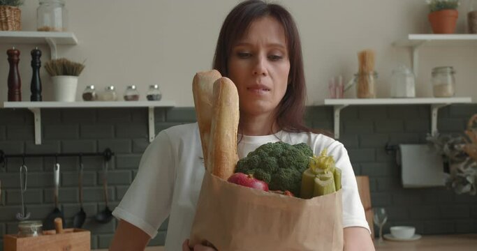 Woman holding paper bag with groceries standing in kitchen looking at camera