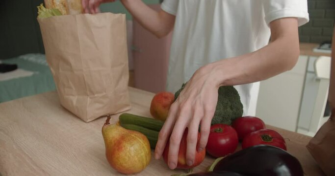 Woman unpacks fresh vegetables and fruits from paper bag with groceries from supermarket delivery on kitchen