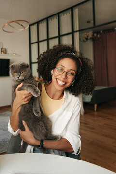 Cute young african girl with snow-white smile holds fluffy cat in her hands. Brunette wears glasses, tank top and shirt at home. Favorite pet concept