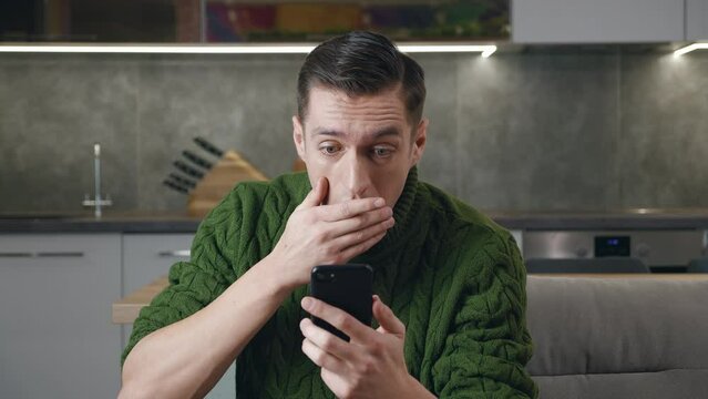 Shocked man reacting on terrible message on smartphone, covers his mouth in fright sitting on sofa at home living room. Unpleasantly surprised and scared male reading bad news on his mobile phone