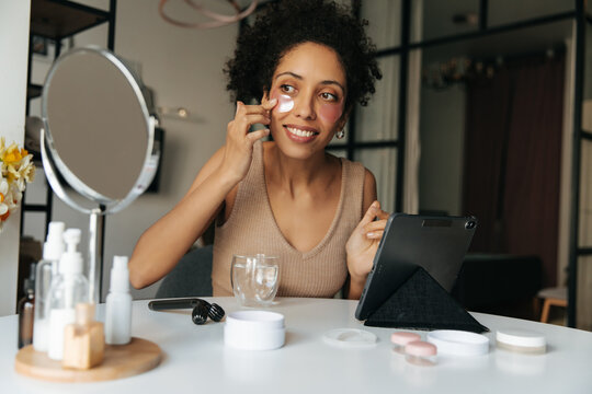 Beautiful young dark-skinned woman applies patches to her face looking in mirror sitting at table. Brunette takes care of the skin, uses cosmetic product. Natural beauty concept