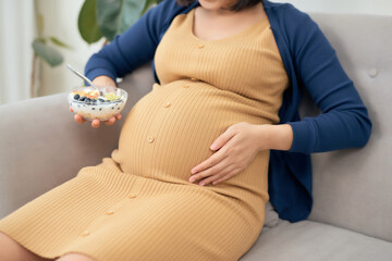 Happy Asian pregnant young woman sitting and eating fruit salad on sofa at home