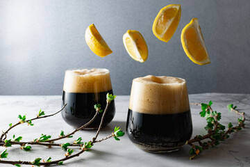 Two low full glasses with a dark carbonated drink with a high foam, beer or kvass, flying lemon slices and a young currant sprig - obrazy, fototapety, plakaty