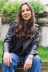 Fototapeta na wymiar Portrait of a young brunette in a leather jacket and jeans. A positive and cheerful young female model is sitting on a bench and posing for the camera