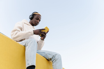 Z generation Black man listening to music with headphones. Casual Sitting on a yellow wall