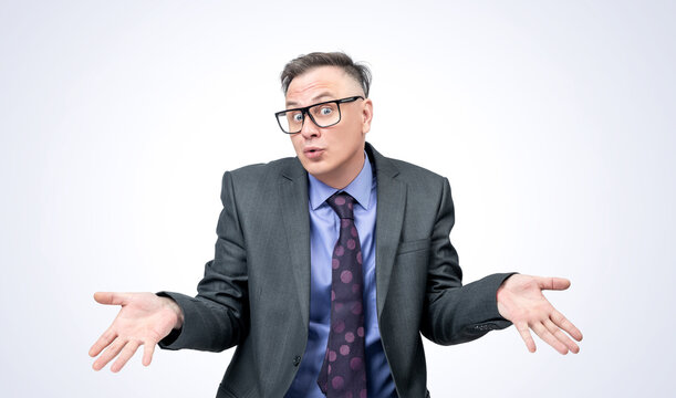 Portrait of a businessman in a dark suit and glasses who shrugs and spreads his arms showing empty palms. On a light background. 