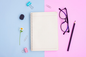 Notebook ,Eyeglasses,Black pencil,Eraser,Paper Clips and White Flower on Pink and Blue Background