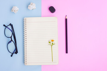 Notebook ,Eyeglasses,Black pencil,Eraser and White Flower on Pink and Blue Background