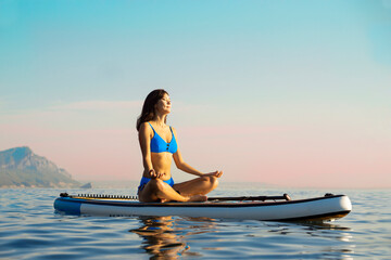 Asian beautiful girl in a blue bikini doing ugha sits in a lotus position with her eyes closed and meditates on a water sports board in the ocean against the background of a sunset. Woman on Sup.