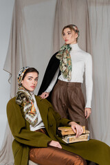 woman in patterned kerchief sitting with vintage books near friend on grey draped background.