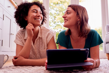 Same Sex Female Couple Or Friends At Home Lying In Lounge Using Digital Tablet And Drinking Coffee
