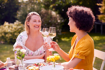 Two Female Friends Sitting Outdoors In Summer Garden At Home Drinking Wine And Eating Meal